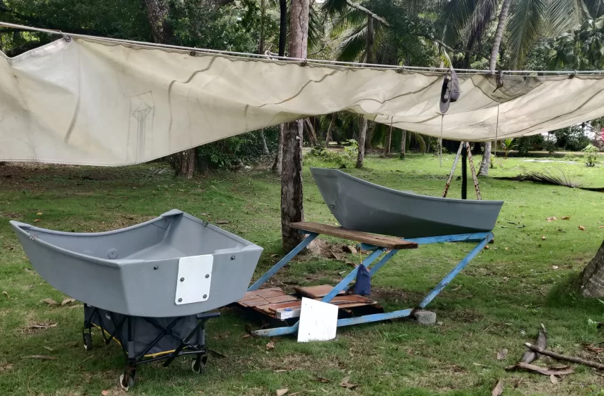two pieces of a small boat are arranged in a makeshift workshop covered by a shade sail beneath palm trees
